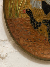 Load image into Gallery viewer, Close-up of an oval decorative wall piece showing a bird in black and gold with green reeds and a brown textured ground, mounted against a white textured wall.

