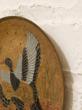 Load image into Gallery viewer, Close-up of an oval decorative wall plaque featuring a painted duck in black, white, and brown tones on a gold background, mounted against a white textured wall.
