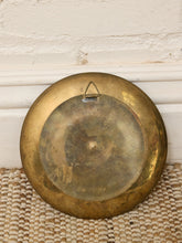 Load image into Gallery viewer, A round vintage brass gong or cymbal with a hanging loop, resting on a woven jute rug against a white brick wall.
