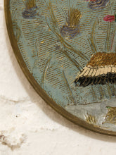 Load image into Gallery viewer, Close-up of an oval decorative wall piece with a gold-toned rim, featuring a textured landscape design in muted blue, gold, beige, and small pink accents against a white plaster wall.
