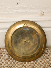 Load image into Gallery viewer, A round vintage brass plate or tray with a small hanging loop at the top, displayed against a white painted brick wall on a woven beige mat.
