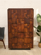 Load image into Gallery viewer, A tall mid-century modern wooden cabinet with an abstract geometric block pattern made of various wood-toned panels, set against a white brick wall with a potted plant and chair partially visible.

