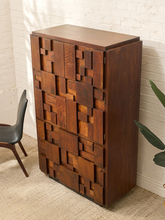 Load image into Gallery viewer, A mid-century modern wooden cabinet with a geometric block pattern on the front, made from multiple shades of brown wood, set against a white brick wall.
