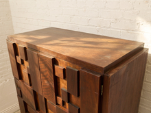 Load image into Gallery viewer, A mid-century modern wooden cabinet with geometric block drawer fronts, shown against a white brick wall with dappled sunlight.
