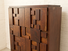 Load image into Gallery viewer, A mid-century modern wooden cabinet with a geometric, block-like design featuring multiple doors and panels in a warm dark brown finish, set against a white brick wall.
