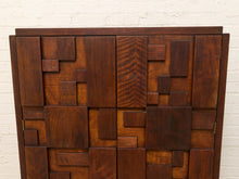 Load image into Gallery viewer, Mid-century modern wooden cabinet with geometric block panel doors in varying shades of dark brown wood, set against a white brick wall.

