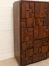 Load image into Gallery viewer, A mid-century modern wooden cabinet with a geometric, block-like pattern of recessed and raised wood panels, finished in a rich dark brown tone, set against a white brick wall and beige carpet.
