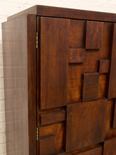 Load image into Gallery viewer, A close-up of a dark brown wooden cabinet featuring a geometric, block-like panel design on the front and side, set against a white brick wall.
