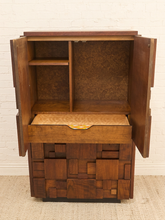 Load image into Gallery viewer, A mid-century modern wooden cabinet with open doors, a pull-out desk surface, and a geometric block-patterned base, shown against a white brick wall and light carpet.
