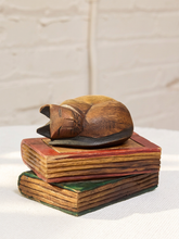 Load image into Gallery viewer, A carved wooden sleeping cat figurine resting on top of a stack of three hand-painted vintage-style books against a white brick wall background.
