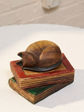 Load image into Gallery viewer, A carved wooden sleeping cat figurine resting on top of two stacked, painted books, displayed on a white textured surface.

