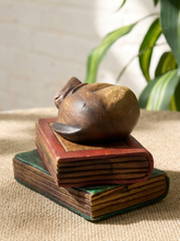 Load image into Gallery viewer, A rustic carved wooden figurine of a sleeping figure, styled on top of two vintage wooden books on a burlap surface with a blurred plant in the background.
