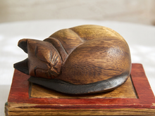 Load image into Gallery viewer, A carved wooden figurine of a sleeping cat resting on a small red and brown wooden base, photographed against a softly blurred light background.
