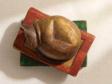 Load image into Gallery viewer, A carved wooden figurine of a sleeping cat resting on a small stack of painted blocks, photographed from above on a textured off-white fabric background.
