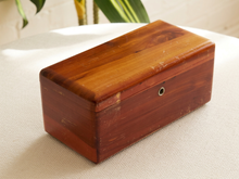 Load image into Gallery viewer, A rectangular vintage wooden box with a warm reddish-brown finish and a small metal keyhole detail on the lid, resting on a textured white fabric surface.
