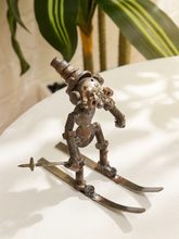 Load image into Gallery viewer, A rustic metal sculpture of a stylized skier made from gears, springs, and rivets, standing on skis with a small sword, displayed on a white textured surface with a blurred plant in the background.
