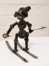 Load image into Gallery viewer, A rustic metal sculpture of a skater figure made from repurposed nuts, bolts, and springs, holding a small metal spear, standing on two metal skis against a white brick wall background.
