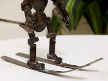 Load image into Gallery viewer, A rustic metal sculpture of a skier made from repurposed nuts, bolts, springs, and metal skis, resting on a white textured surface with a blurred green plant in the background.
