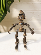 Load image into Gallery viewer, A small handmade metal robot figurine made from screws, nuts, bolts, and springs, standing on skis. The figurine has a weathered brass and steel finish and is photographed on a white surface with a white brick wall and a green plant leaf in the background.
