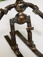 Load image into Gallery viewer, A handmade sculpture of a small robot or mechanical figure made from rusty metal bolts, nuts, and threaded rods, standing on two long metal bars against a plain light background.

