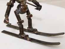 Load image into Gallery viewer, A close-up of a rustic metal sculpture of a skier, constructed from repurposed bicycle components including threaded springs, rusty bolts, and dark metal skis, set against a plain white textured background.
