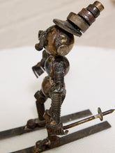 Load image into Gallery viewer, A rustic metal sculpture of a stylized skier made from repurposed metal parts, including a large nut for a hat, standing on metal skis against a light background.
