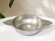 Load image into Gallery viewer, A vintage silver metal bowl with decorative leaf-shaped side handles, resting on a white textured tablecloth against a white brick wall background.
