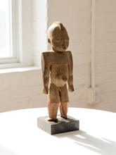 Load image into Gallery viewer, A hand-carved wooden figurine of a standing nude human form on a dark rectangular base, displayed on a white table in front of a white brick wall and window.
