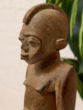 Load image into Gallery viewer, A close-up of a hand-carved wooden sculpture of a stylized human head and upper torso, shown in profile against a softly blurred background with a light wall and green leaves.
