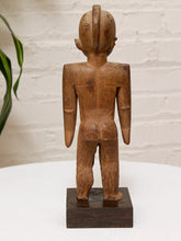 Load image into Gallery viewer, A hand-carved wooden figurine of a standing human figure, shown from the back on a dark rectangular base against a white brick wall background.
