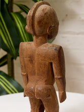 Load image into Gallery viewer, A hand-carved wooden figurine of a nude human figure, shown from the back, featuring detailed wood grain and a stylized hairstyle, set against a background of a potted plant and a light-colored wall.
