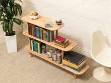 Load image into Gallery viewer, A three-tier wooden bookshelf with rounded edges and tapered legs, styled with books, a fruit bowl, and decorative items in a bright room with a white brick wall, a potted plant, and a modern chair.
