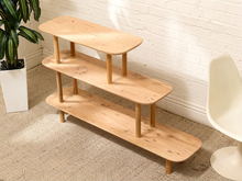 Load image into Gallery viewer, A three-tier wooden shelf with rounded rectangular shelves and tapered legs, shown in a styled room setting with a white brick wall, beige carpet, a potted plant, and a white chair.

