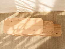 Load image into Gallery viewer, A minimalist light natural wood bench with rounded rectangular legs, photographed from a top-down angle on a textured beige rug with dappled sunlight and shadows.
