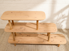 Load image into Gallery viewer, A three-tier wooden shelving unit made of light natural wood, set against a white brick wall and a textured beige rug.
