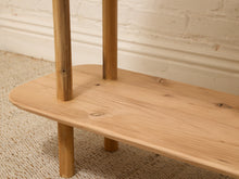 Load image into Gallery viewer, Close-up of a light natural wood furniture piece with a rounded rectangular shelf or bench top and cylindrical legs, shown against a white brick wall and beige carpet.
