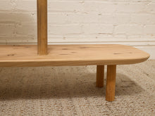 Load image into Gallery viewer, A close-up of a light natural wood furniture piece, showing a shelf or bench section with a vertical post and two legs, set on a woven rug against a white brick wall.

