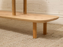 Load image into Gallery viewer, A close-up of a light natural wood bench with cylindrical legs, positioned on a woven beige rug against a white painted brick wall.
