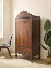 Load image into Gallery viewer, A dark brown carved wooden armoire cabinet with ornate details, set in a room with a white brick wall, a black chair, and a potted plant.
