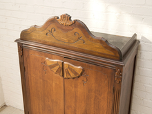 Load image into Gallery viewer, An antique wooden cabinet with ornate carved details and a scalloped top panel, photographed against a white painted brick wall.
