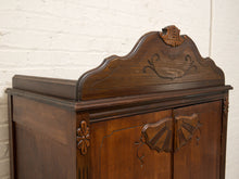 Load image into Gallery viewer, An antique dark brown wooden cabinet with ornate carved details, featuring a decorative arched top panel and two doors with fan-shaped inlays, set against a white brick wall.
