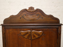 Load image into Gallery viewer, A close-up of a dark brown wooden cabinet featuring ornate hand-carved details, including a fan-shaped motif on the doors and a decorative crest on the arched top panel, set against a white brick wall background.
