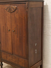 Load image into Gallery viewer, A close-up of a dark brown wooden cabinet with ornate carved details on the upper door, vertical panel accents, and small round knobs, shown against a white brick wall.
