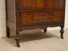 Load image into Gallery viewer, A close-up of a dark brown wooden chest of drawers with turned legs, positioned against a white brick wall and beige carpet.
