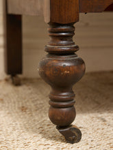 Load image into Gallery viewer, Close-up of a dark brown wooden table leg with a turned spindle design and a small caster wheel at the bottom, resting on a woven beige rug.
