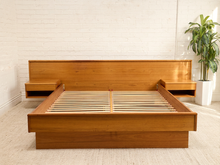 Load image into Gallery viewer, A modern low-profile platform bed frame made of warm medium-brown wood, shown in a minimalist bedroom setting with a white brick wall, light beige carpet, and a potted green plant on the right.
