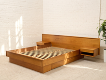 Load image into Gallery viewer, A mid-century modern platform bed frame made of warm medium-brown wood, featuring a matching headboard, floating side nightstands, and a slatted base, set in a bright room with a white brick wall and light beige carpet.

