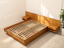 Load image into Gallery viewer, A low-profile platform bed frame made of warm medium-brown wood, featuring a matching headboard and two floating side tables, shown in a bright room with white brick walls and light-colored flooring.
