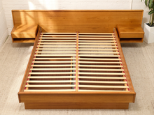Load image into Gallery viewer, A modern wooden platform bed frame with an integrated headboard and side nightstands, shown with the slatted base extended. The bed is made of warm medium-brown wood and is photographed in a bright room with a light beige rug and white brick wall.
