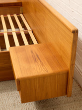 Load image into Gallery viewer, Close-up of a wooden bed frame corner with a pull-out storage drawer, showing warm medium-brown wood grain, slatted support bars, and a white brick wall background.
